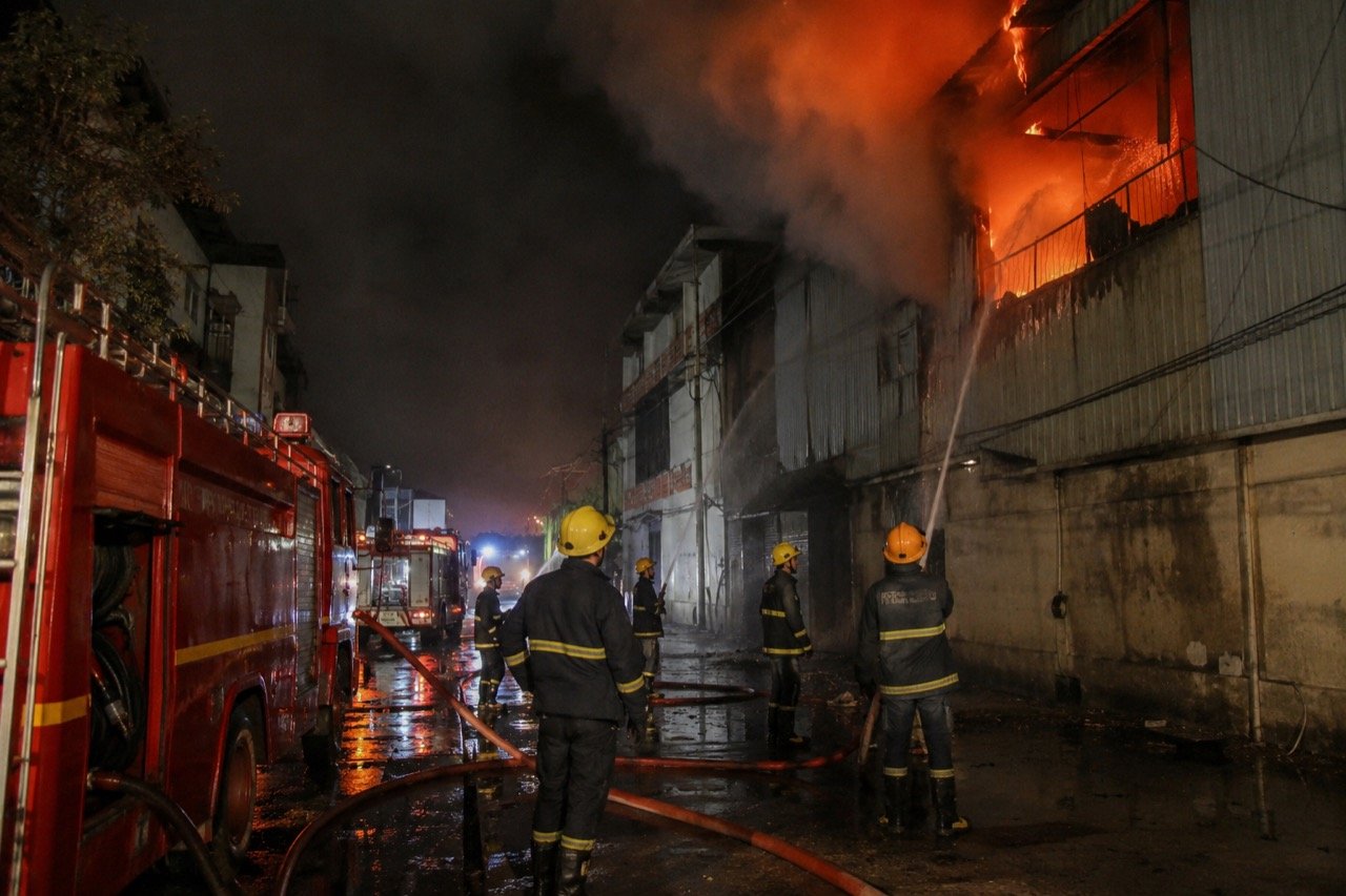 Fire engines with flashing lights responding to smoke rising from a factory building in a narrow Delhi industrial lane at night