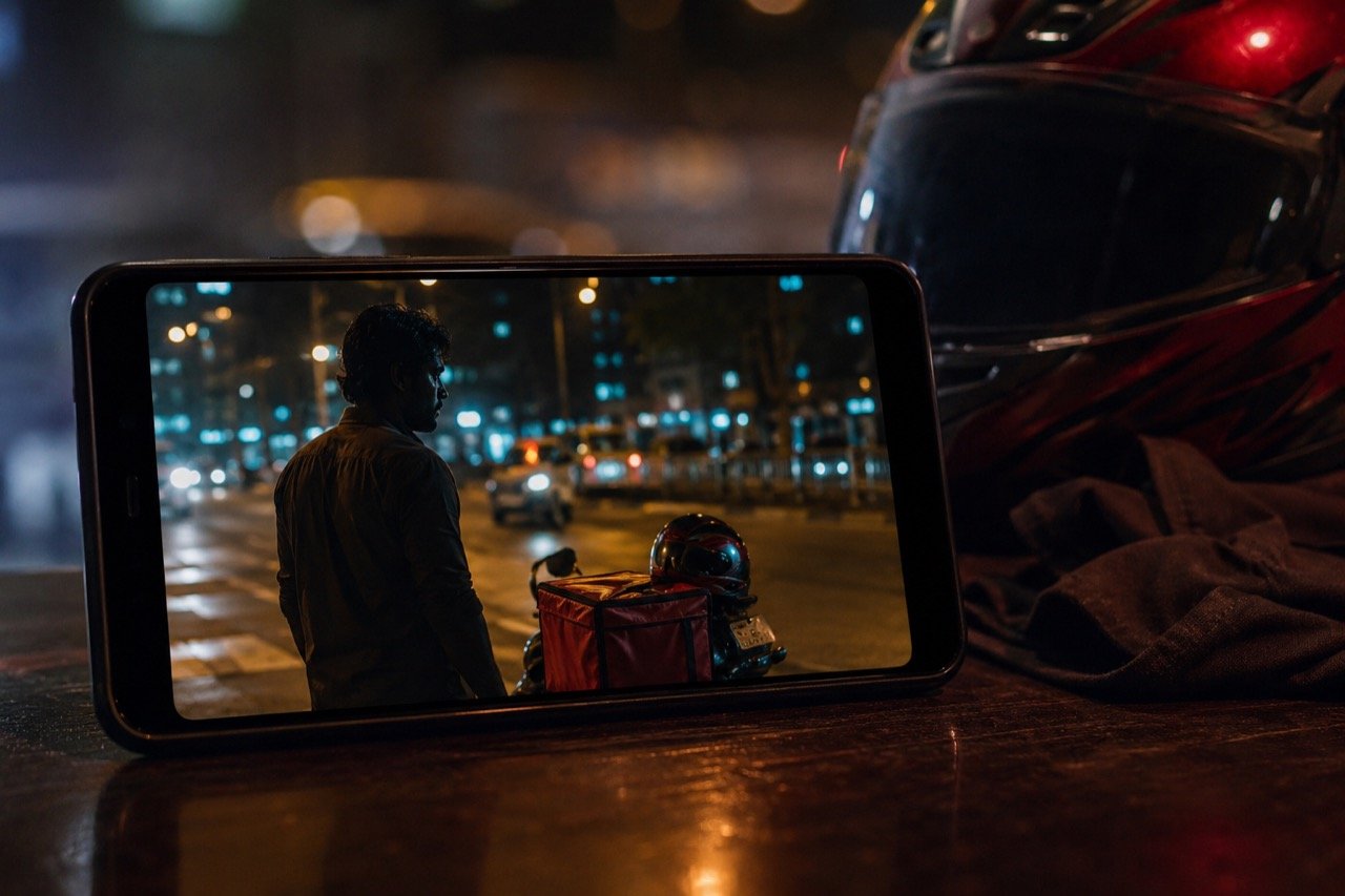 A delivery rider on a motorbike speeding through neon-lit city streets at night, evoking the suspenseful mood of a Tamil action thriller
