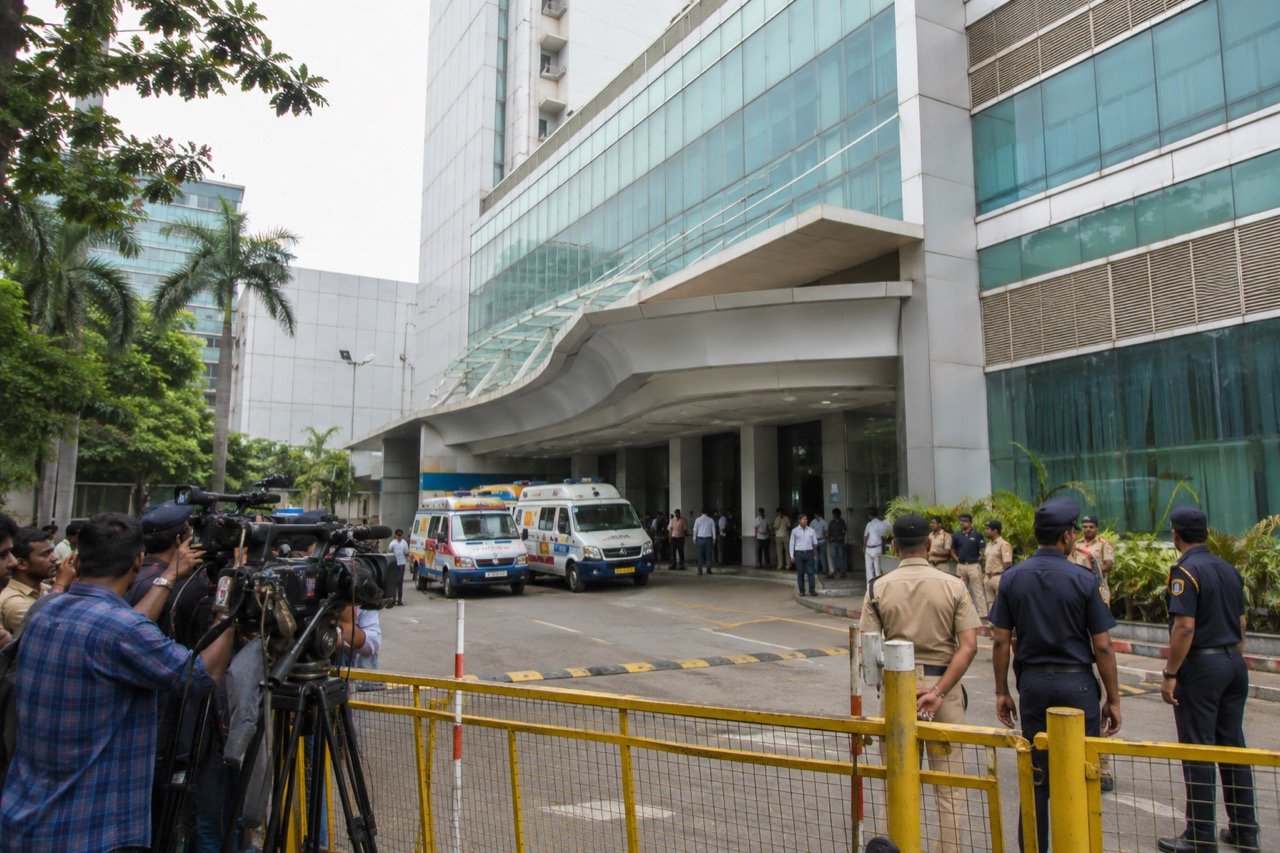 Exterior of a Mumbai hospital building at dusk with media vans and onlookers gathered near the entrance