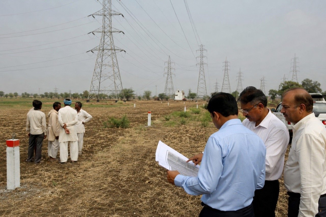 High-voltage transmission towers and power lines crossing green farmland in Gujarat under a bright sky, with a farmer's field in the foreground