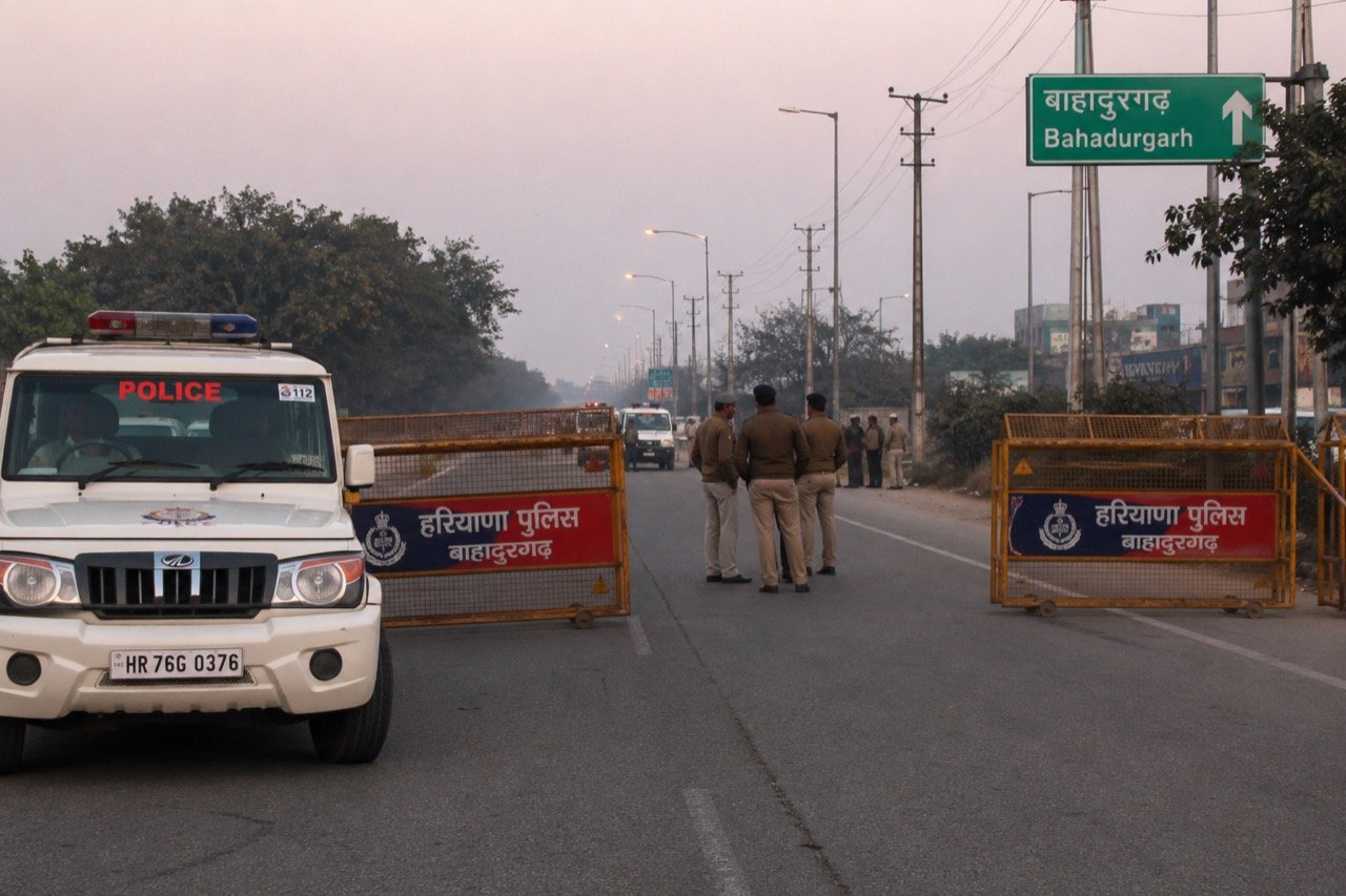 Police vehicles and armed officers cordoning off an encounter site on a road in Bahadurgarh, Haryana, at dusk with flashing lights