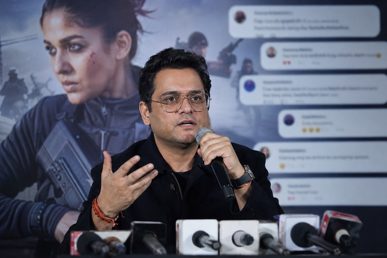 Karan Johar speaking at a film industry event with a poster-style backdrop suggesting the spy-action film Alpha behind him