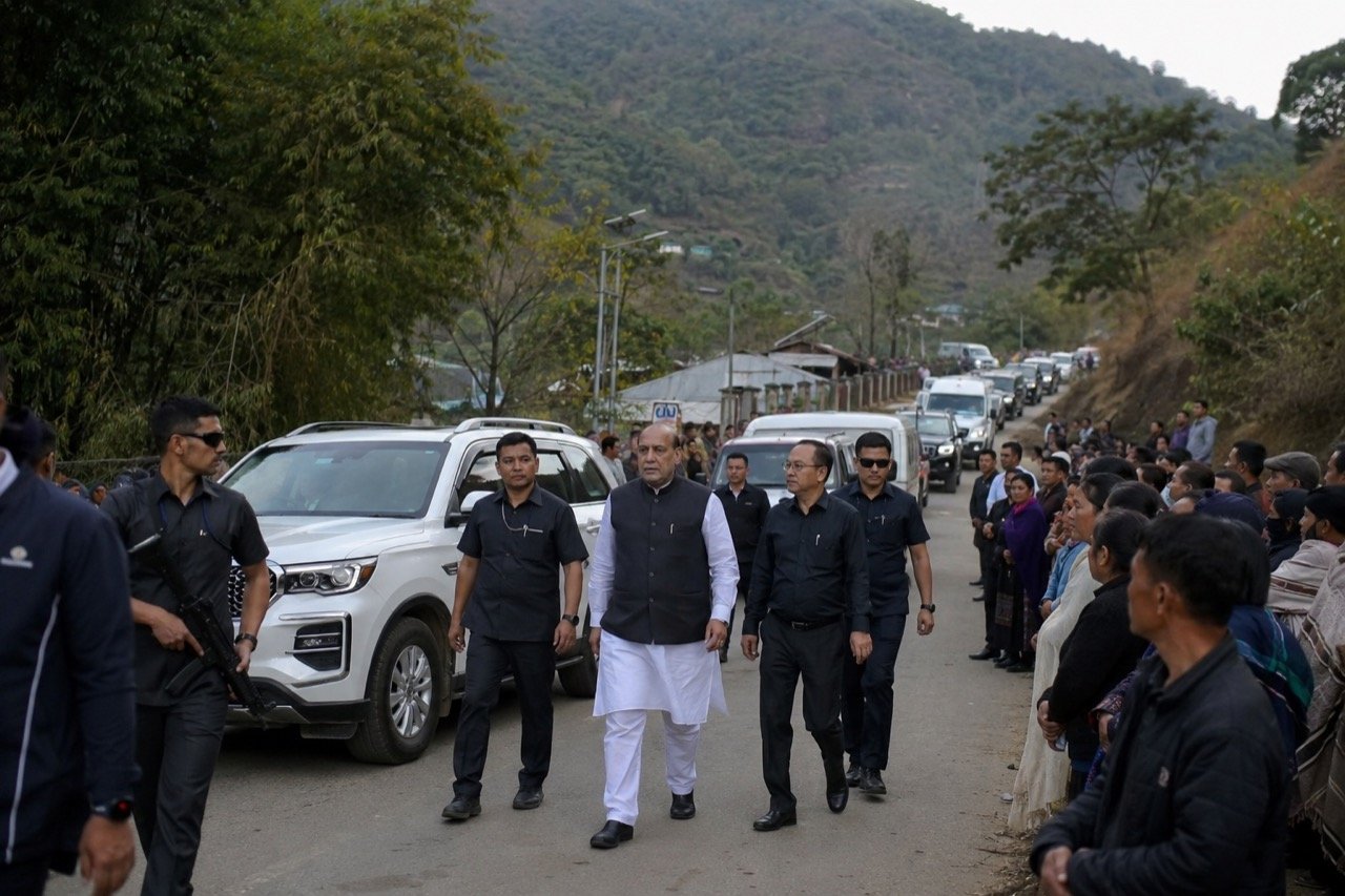 A convoy of official vehicles moving along a quiet hill road in Churachandpur, Manipur, with misty hills and a solemn gathering in the distance