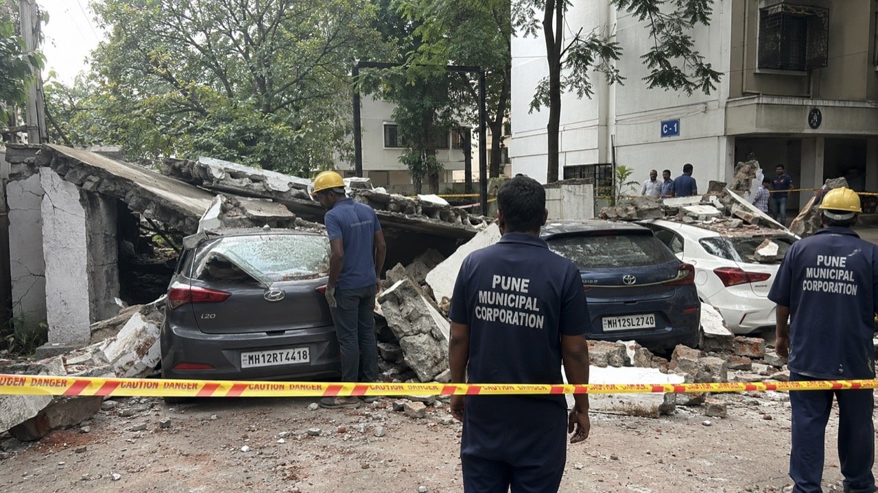 A collapsed brick compound wall in a rain-soaked Pune lane, rubble crushing parked two-wheelers and cars as onlookers gather under grey monsoon skies